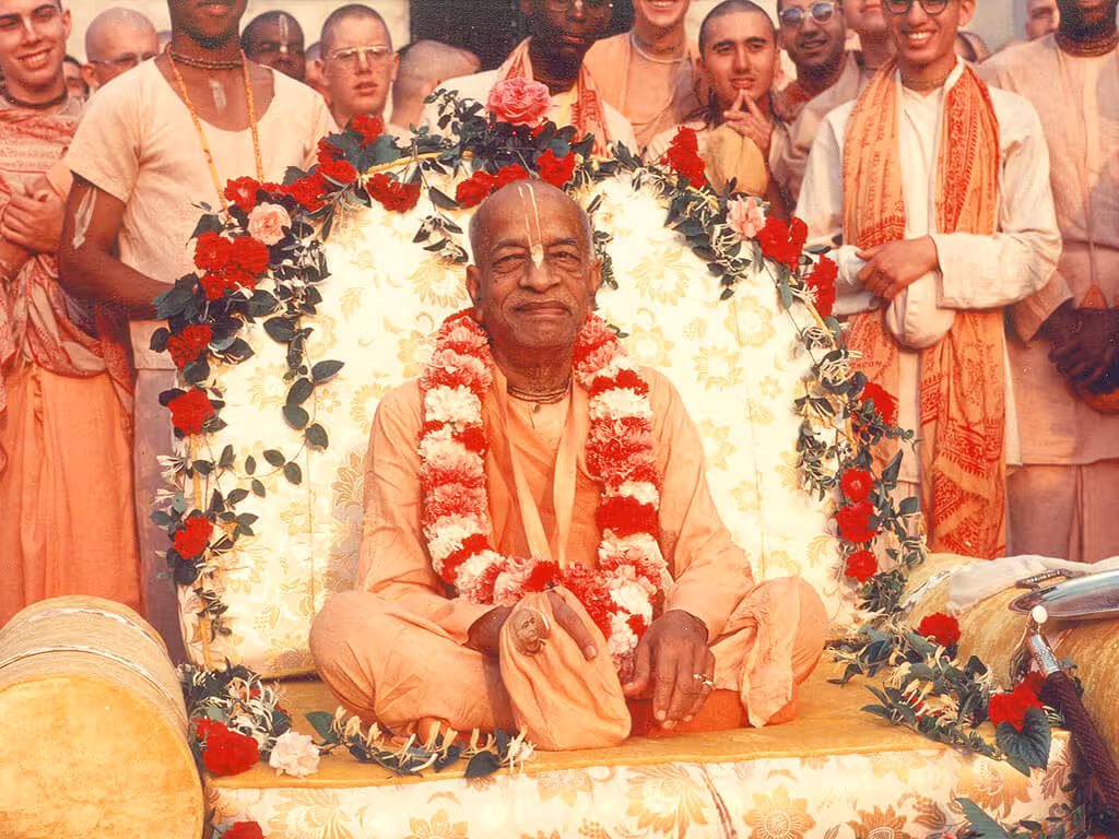 His Divine Grace A.C. Bhaktivedanta Swami Prabhupada seated on a vyasasana, adorned with flower garlands, surrounded by smiling disciples